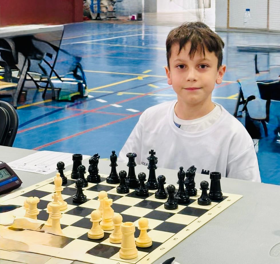 Young boy in a white shirt sits at a chessboard in a gymnasium, ready to play with a timer nearby.