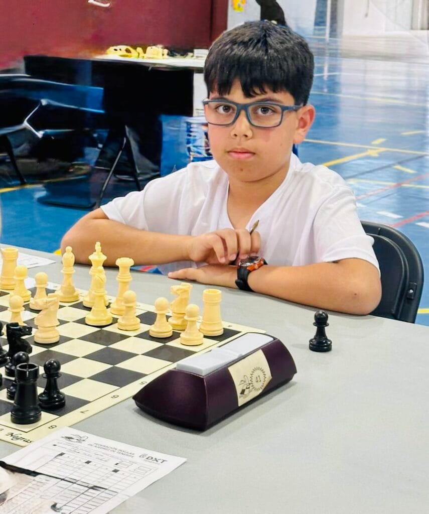 A young boy with glasses sits at a chessboard during a tournament, preparing a move.