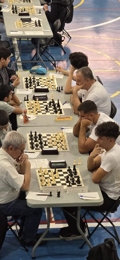 Several players compete in a chess tournament at folding tables in a gym, with clocks and score sheets on the boards.