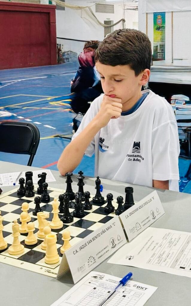 Young boy studies a chessboard during a tournament, hand on chin, wearing a white shirt with logos, blue gym floor in background.