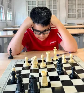 Young boy in a red Adidas shirt and glasses leans on the desk, concentrating on a chessboard in a classroom.