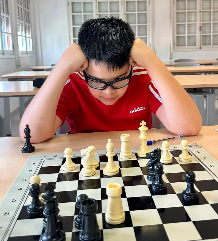 Young boy in a red Adidas shirt and glasses leans on the desk, concentrating on a chessboard in a classroom.