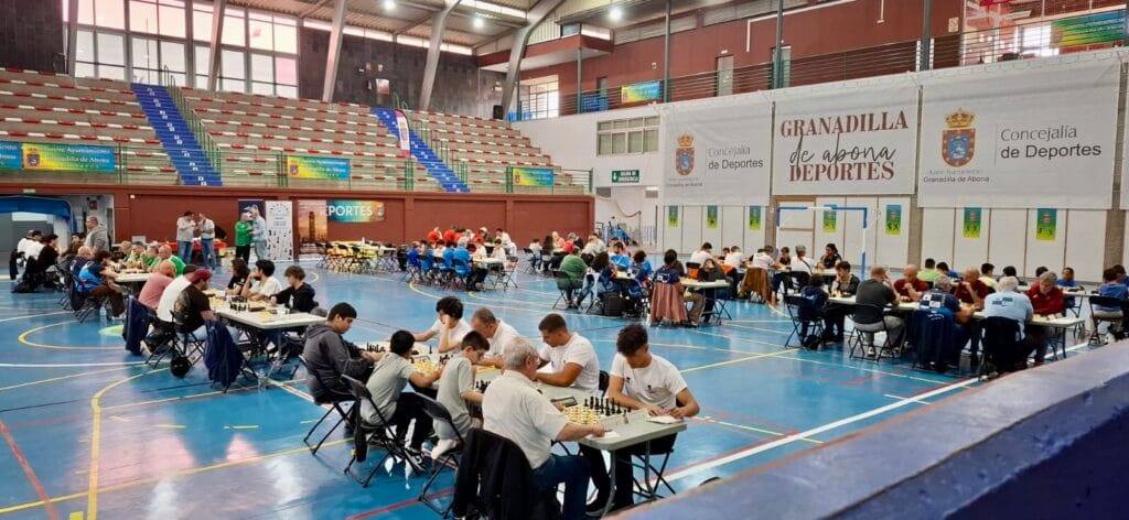 Indoor sports hall hosting a chess tournament with many tables and players focused on games, banners on the walls, and spectators in the background.