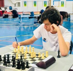 Young man in a white shirt sits at a chessboard in a gym, writing on a score sheet while resting his chin on his hand.