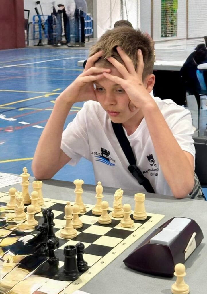 Boy at a chess tournament with hands on his head, concentrating over the board in a gymnasium.