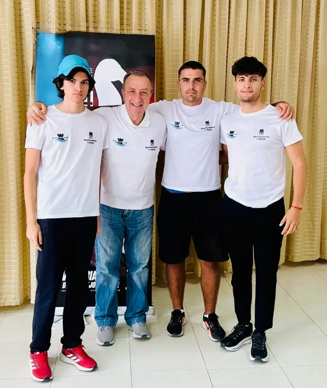 Four men in matching white logo shirts pose with arms around each other indoors, standing in front of a beige curtain.