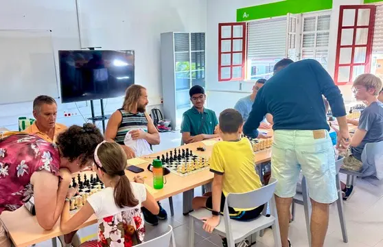 Group of children and adults seated at tables, playing chess in a bright classroom; a man stands and watches as players think.