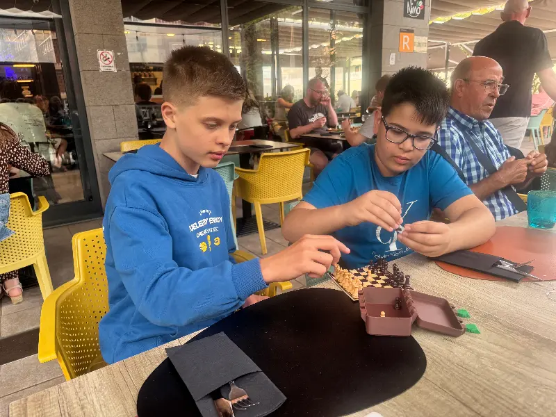 Two boys in blue shirts building a chess set at an outdoor cafe; yellow chairs around, a man sits nearby.