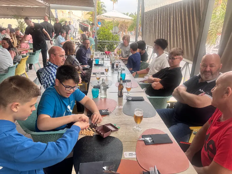 Group of diners at an outdoor patio, two boys in blue shirts playing chess at a long table while others chat and drink.