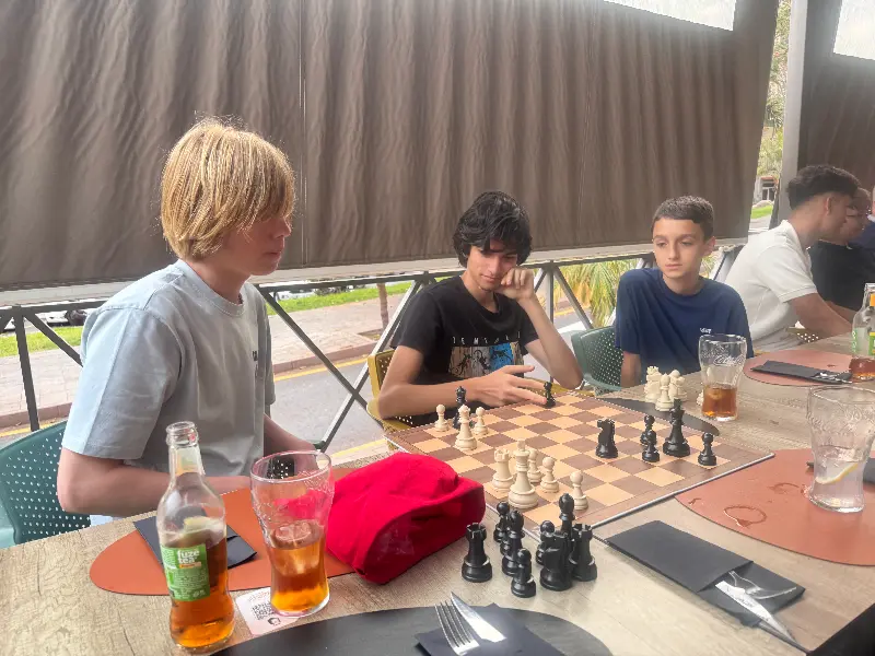 Group of young people playing chess at an outdoor table, with drinks and a red bag nearby.