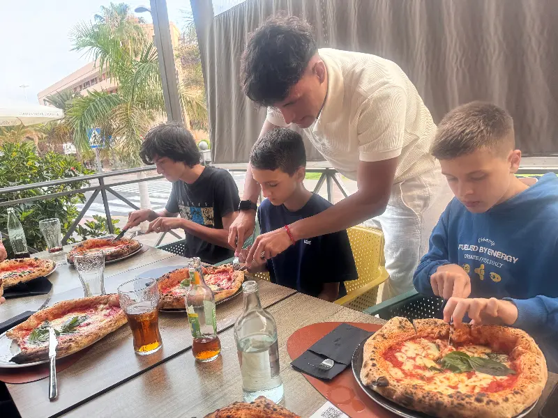 Man helps two kids and a teen prepare pizzas at an outdoor table with drinks and bottles nearby