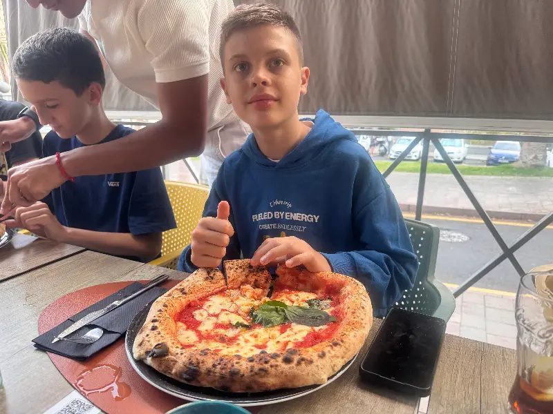 Boy in a blue hoodie gives a thumbs-up while eating a Margherita pizza at an outdoor cafe table.