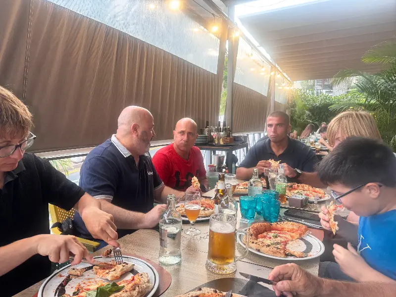 Group of people dining at an outdoor restaurant, sharing pizzas and drinks around a long table.