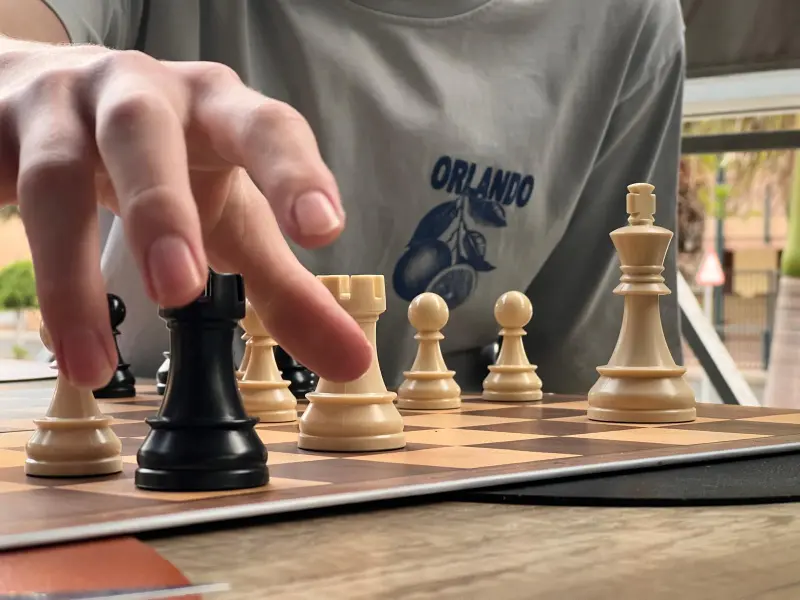 Close-up of a hand moving a black chess piece on a wooden chessboard outdoors, with a gray shirt reading 'Orlando' in the background.