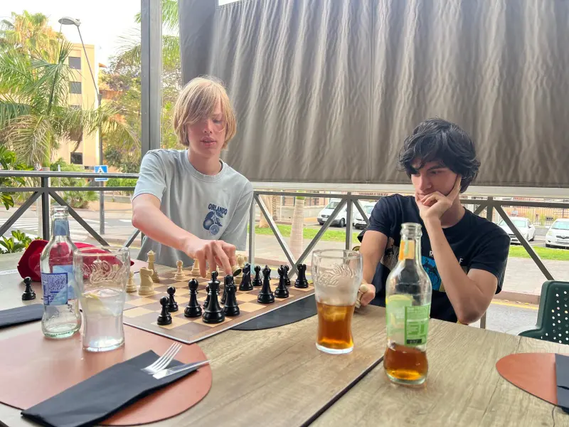Two young men play chess at an outdoor patio, with beers and water bottles on the table nearby.