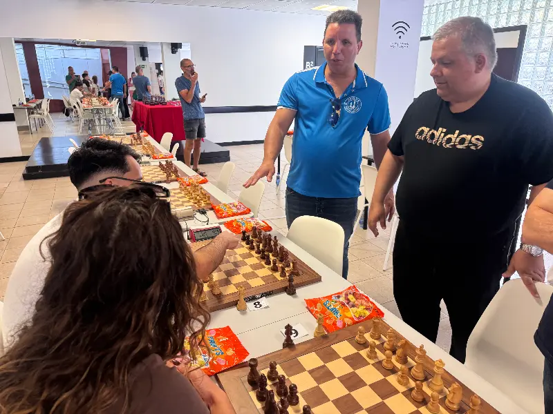 People play chess at a tournament in a bright room; players sit at tables with boards and chips nearby, while two spectators observe the game.